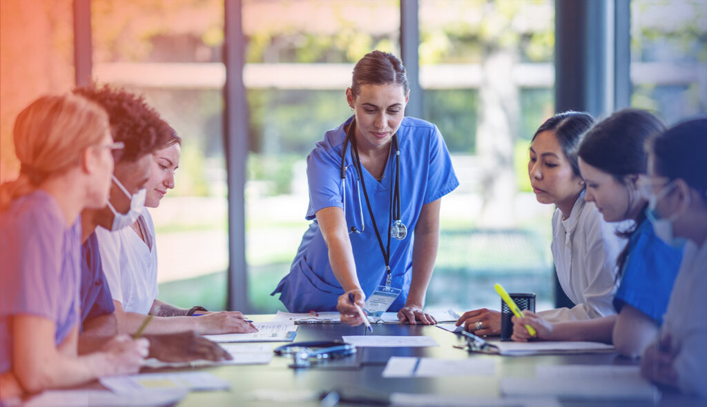 Nurse stands before seated colleagues and points to a document on the table.