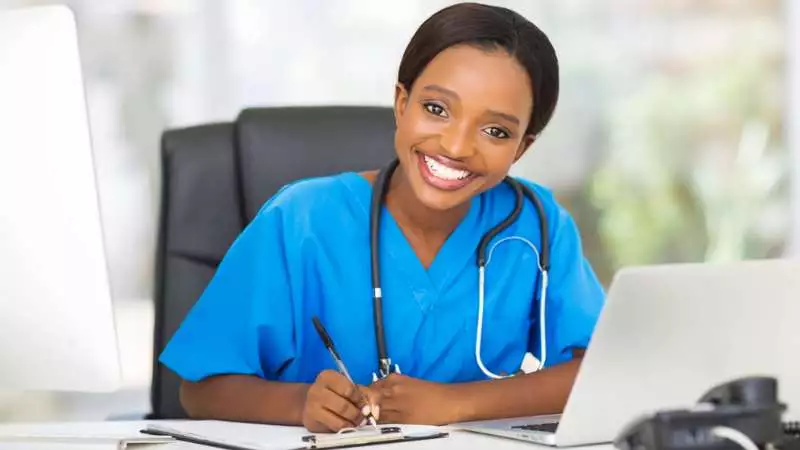 A nurse wearing scrubs and a stethoscope smiles at the camera in the middle of taking notes on a clipboard.