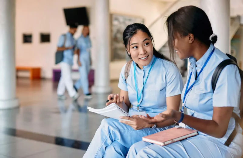 Two student nurses compare notes in a hospital hallway.