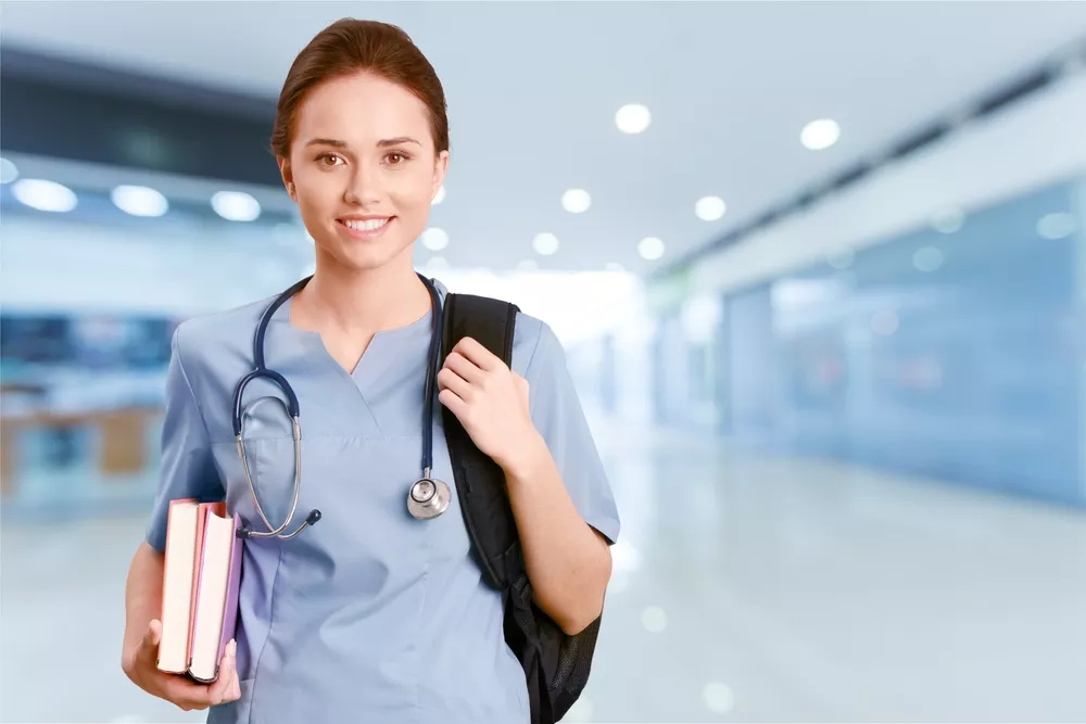 A nurse in scrubs wears a stethoscope and a backpack and carries two books.