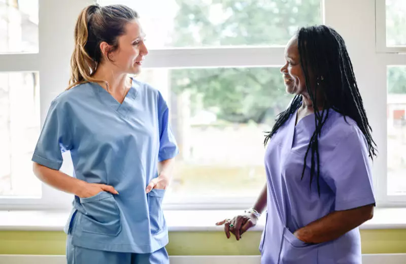 Two nurses in scrubs speak with each other in a healthcare facility.