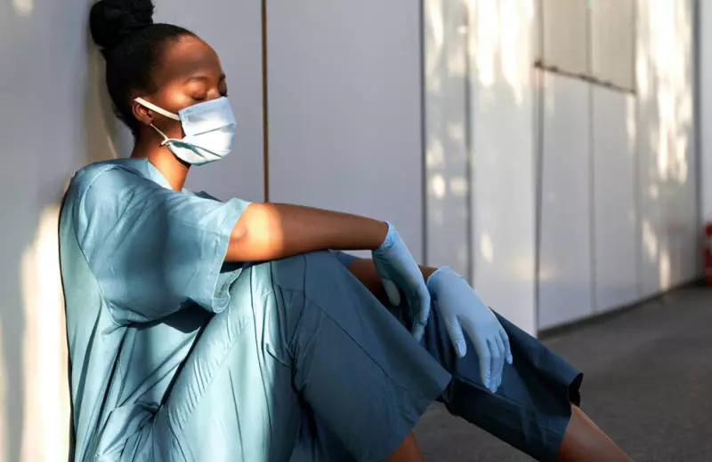 A nurse wearing scrubs and a face mask sits on the ground against a wall with her eyes closed.