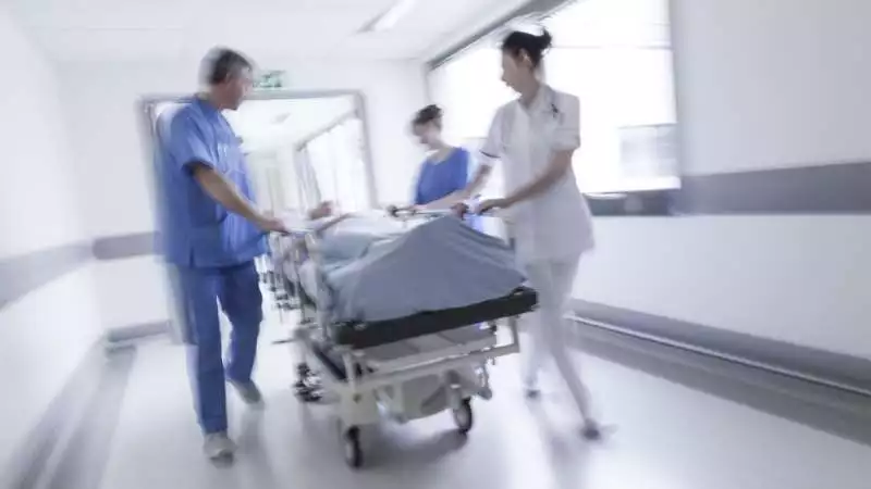 Three nurses wheel a patient on a stretcher down the hallway of a healthcare facility.