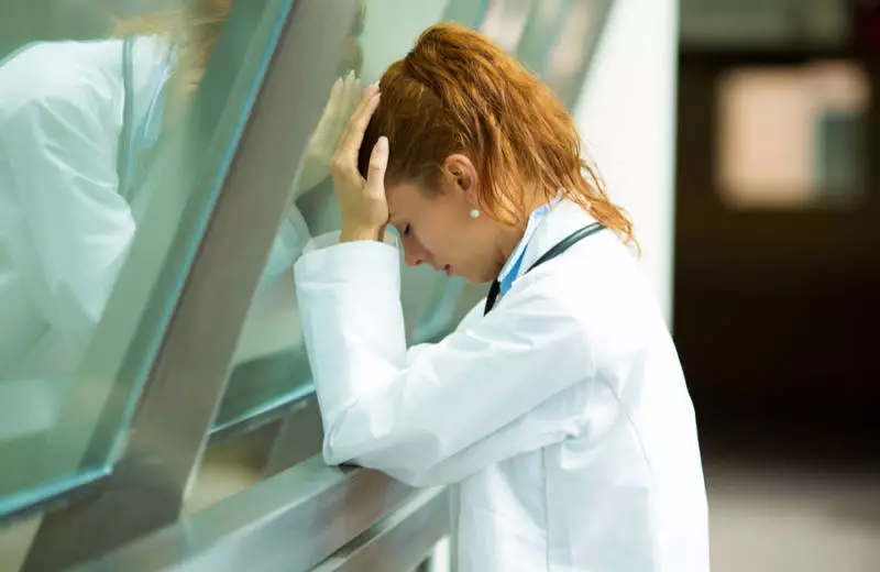 A nurse wearing a white coat closes her eyes and holds her head in her hands.