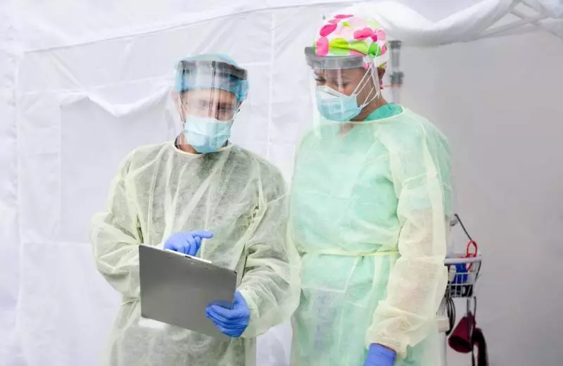 Two nurses in full protective gear study information on a clipboard.