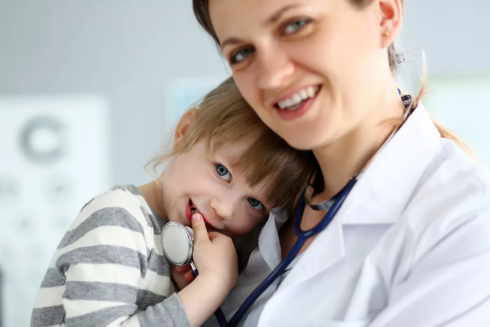 A family nurse practitioner holds a young patient in her arms who plays with her stethoscope.