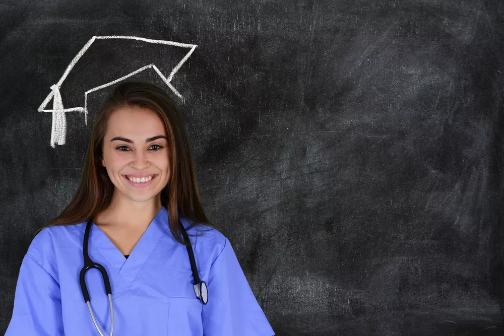 A nursing professional wearing a stethoscope and scrubs stands in front of a chalkboard where there is a graduation cap drawn above her head.