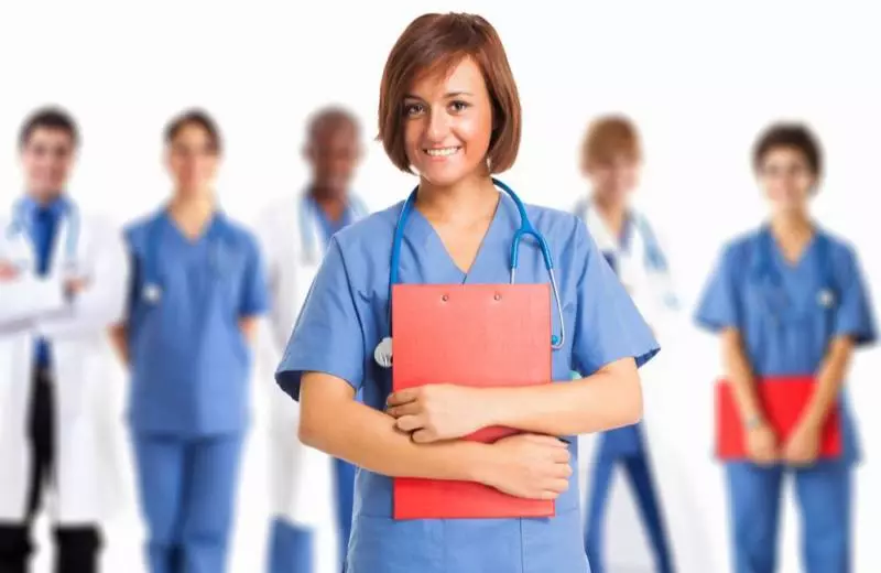 A nursing professional holding a red clipboard smiles at the camera with a group of healthcare professionals in the background.