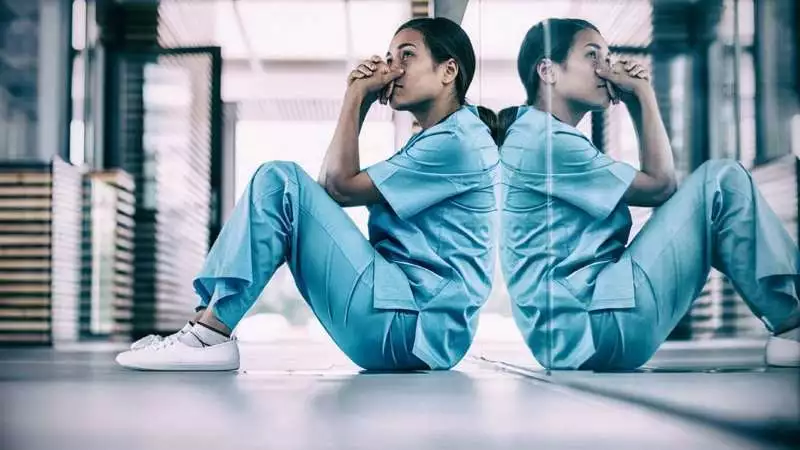 A nurse in scrubs sits on the floor with her head in her hands.