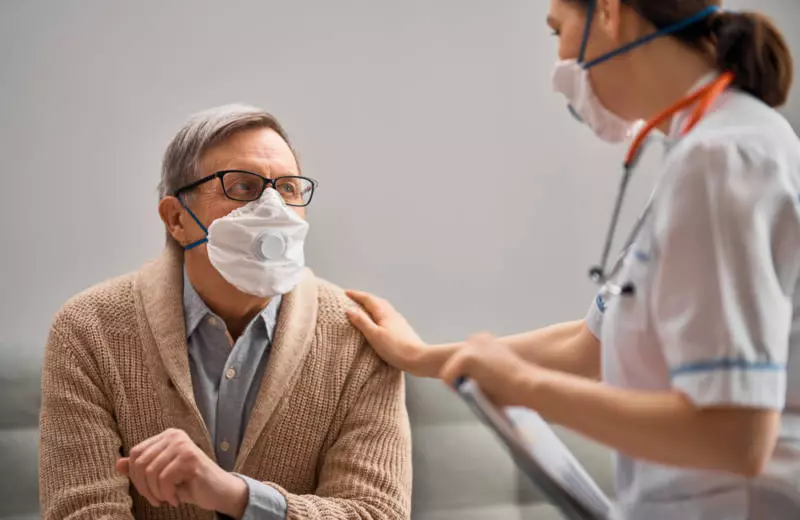 A nurse wearing a mask gently touches an older patient on the shoulder.