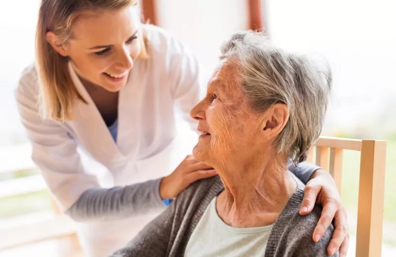 A nurse places her hands gently on an older patient's shoulders and smiles at her.