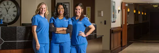 Three nurses in blue scrubs stand in the hallway of a health care facility.