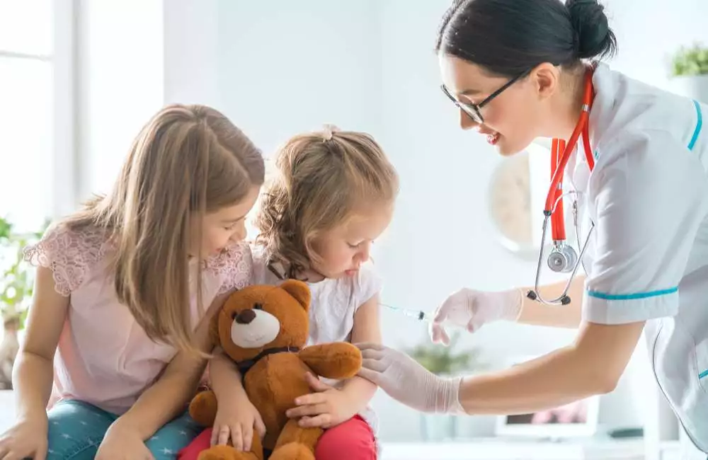 A nurse practitioner prepares a shot for a young patient who sits beside her sister.