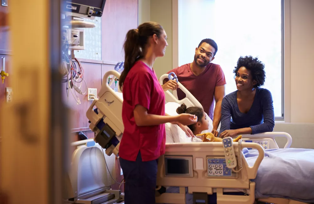 A pediatric nurse practitioner stands beside a young patient's hospital bed speaking to the patient's parents.