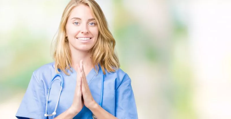 A nurse practitioner smiles at the camera with her palms folded in prayer.