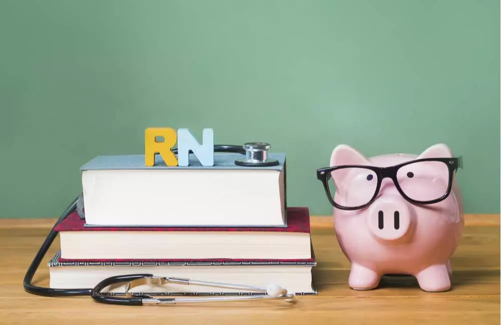 The letters "RN" and a stethoscope sit on top of a stack of books, beside a piggy bank wearing glasses.