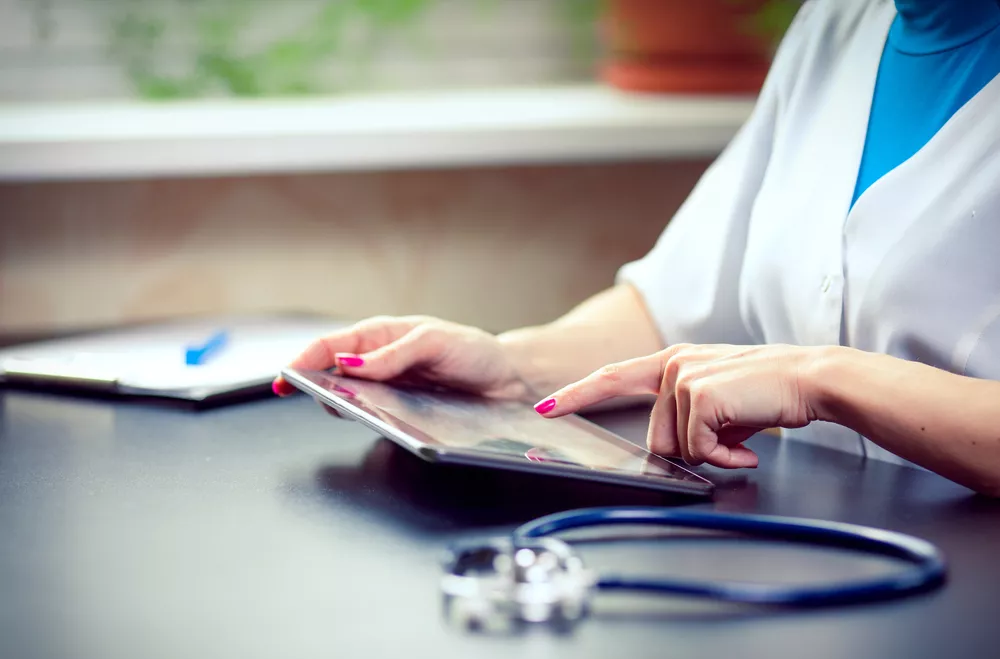 A closeup of a nurse's hands using a tablet on a table, beside a stethoscope.