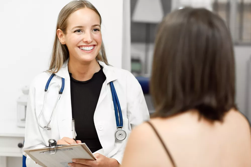 A nurse practitioner wearing a stethoscope carries a clipboard and speaks to a patient.