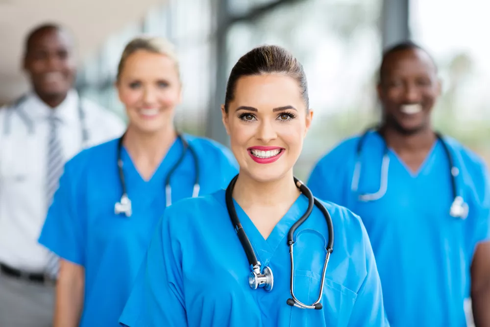 A group of nurses in blue scrubs with stethoscopes around their neck smile at the camera.