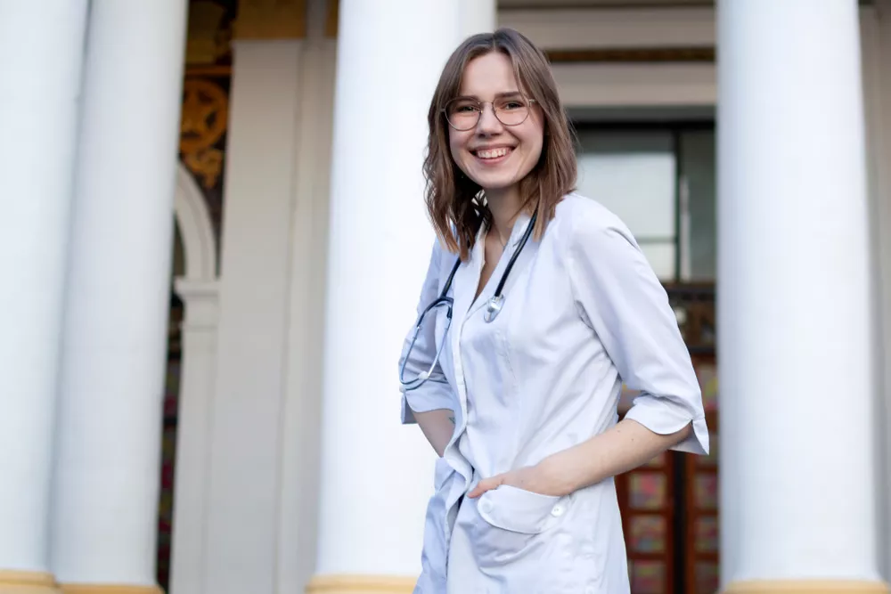 A nurse practitioner wearing a white coat and stethoscope stands in front of a healthcare facility.