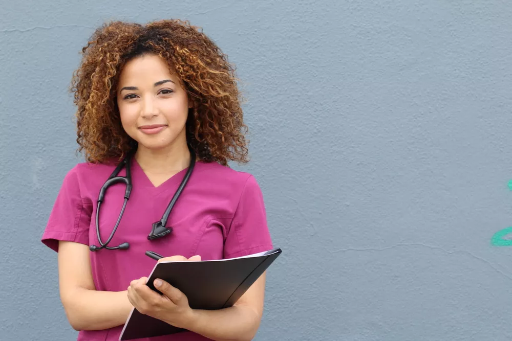 A nurse wearing scrubs and a stethoscope holds a clipboard and smiles at the camera.