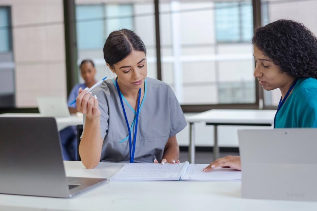 Two nurses seated at laptop computers review hardcopy documents together.